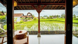 A wooden structure with a tiled floor, a table, and chairs, overlooking a rice field and houses. - Puri Prana Villa (Ubud)