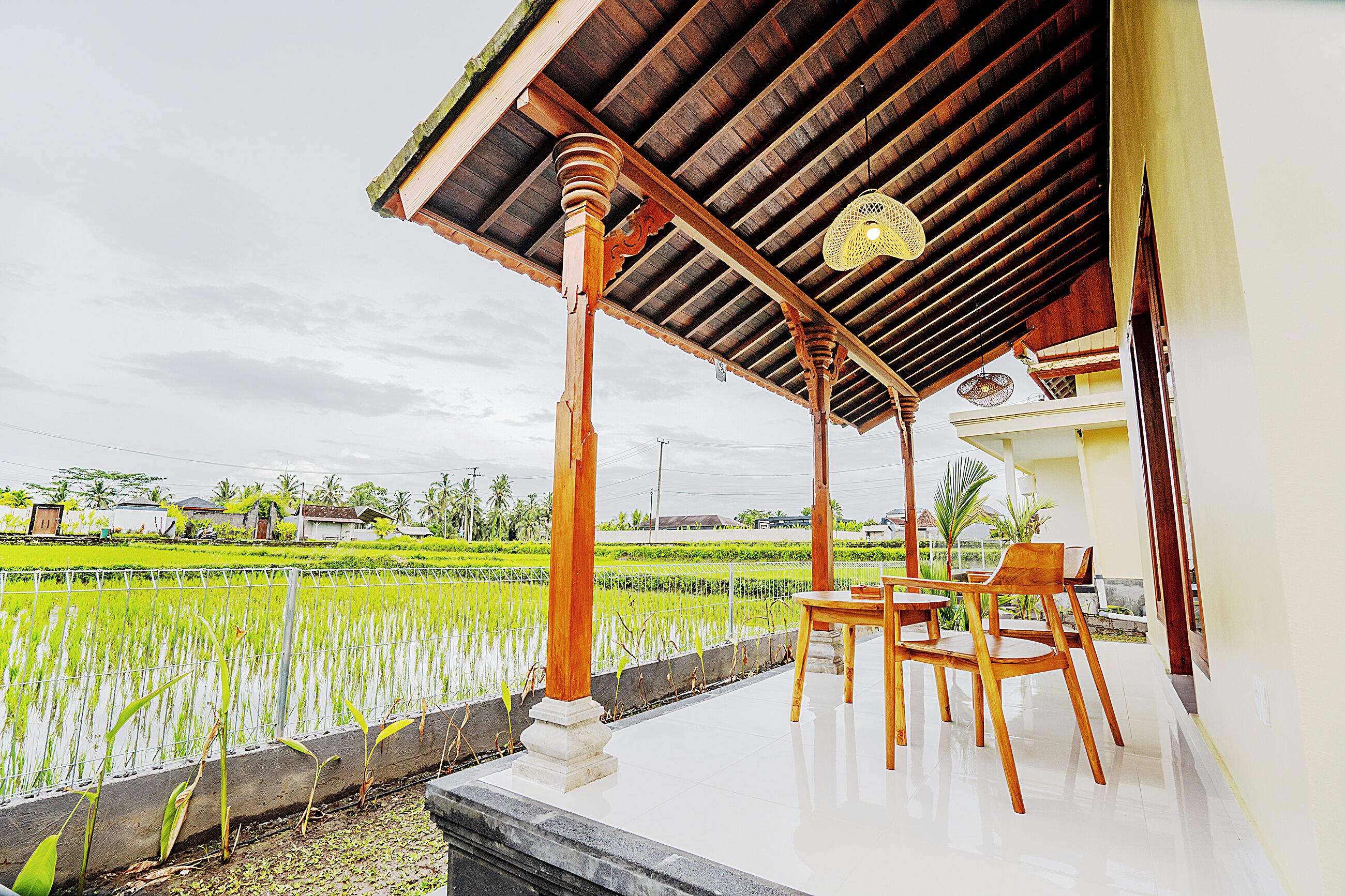 A covered porch with wooden pillars, a table, and chairs, overlooking a rice field.