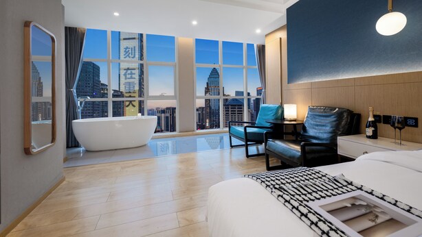 Habitación de hotel moderna con una bañera grande, una cama, un escritorio y una botella de champán, y vistas a la ciudad a través de amplios ventanales.