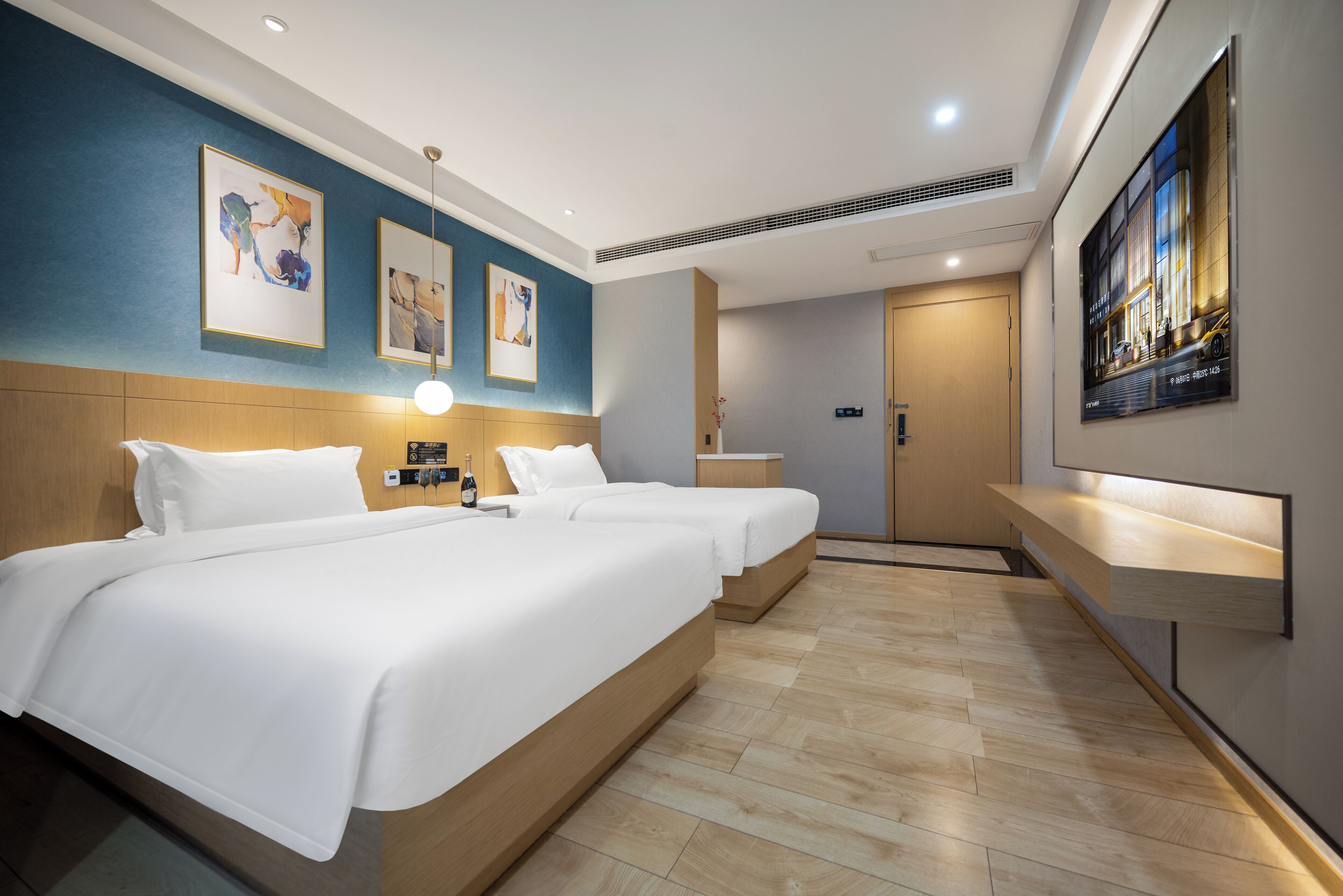 A hotel room with two beds, a wooden headboard, a bedside table with a lamp, a wall-mounted TV, and artwork.