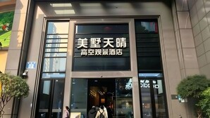 Entrada de un hotel con fachada de vidrio moderna, una placa con caracteres chinos y una vista despejada del interior a través del vidrio.