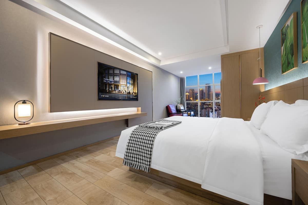 Une chambre d’hôtel moderne dotée d’un grand lit, d’un téléviseur fixé au mur, d’un bureau avec une chaise et offrant une vue sur la ville par la fenêtre.