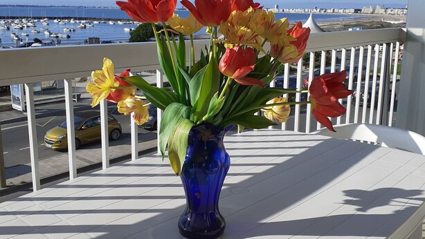 En balkon med en vase med tulipaner, en hvid stol og udsigt over havet og både.