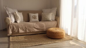 Ein beiges Sofa mit gemusterten Kissen, ein gewebter Hocker und ein gewebter Teppich in einem Raum mit durchsichtigen Vorhängen.