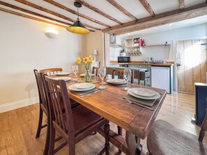 Un coin repas avec une table en bois dressée pour quatre personnes, un vase rempli de fleurs et, au fond, une cuisine.
