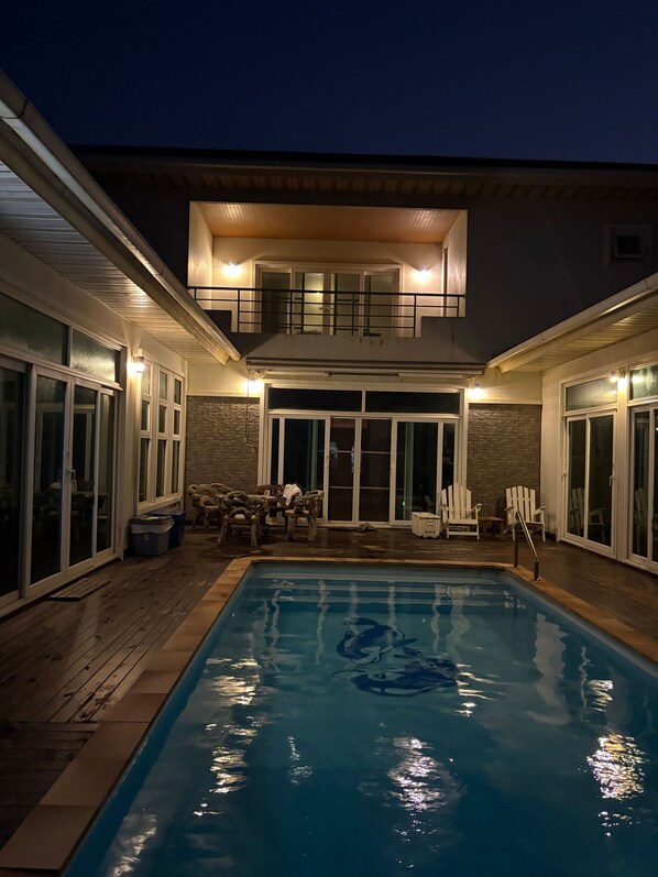 A swimming pool with a wooden deck, a stone wall, and a covered patio area with chairs.