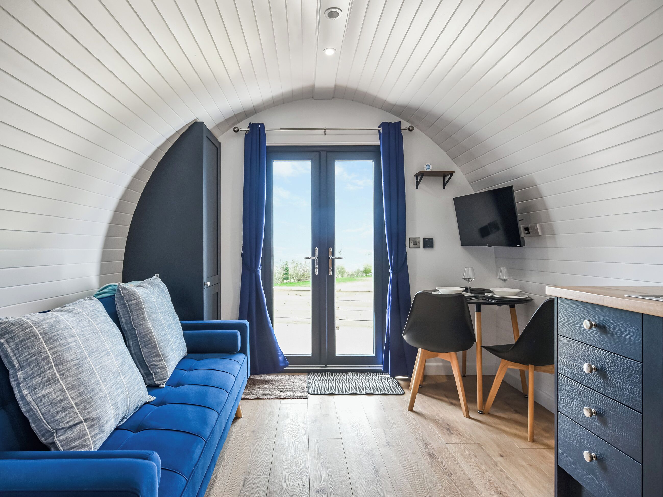 A compact room with a blue sofa, a small desk with a chair, a TV mounted on the wall, and a door leading outside.