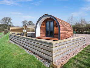 Eine moderne Holzblockhütte mit gewölbtem Dach, einer Terrassenfläche mit Sitzgelegenheit und einem Kiesweg.