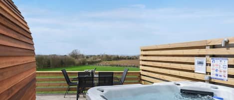 A hot tub with a wooden deck, a table with chairs, and a view of a green field.
