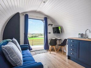 Ein gemütliches Zimmer mit einem blauen Sofa, einer kleinen Essecke mit zwei Stühlen, einem an der Wand montierten Fernseher und Blick ins Freie.