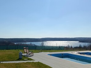 A swimming pool with a view of a lake, surrounded by a fenced area and a grassy lawn.