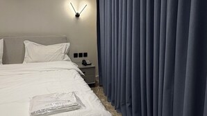 A modern hotel room with a neatly made bed, a wall-mounted light, and a large window with blue curtains. - 009 tara-JRE Jabal Al Lweibdeh with stunning view (Amman)