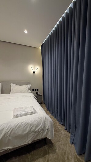 A modern hotel room with a neatly made bed, a wall-mounted light, and a large window with blue curtains. - 009 tara-JRE Jabal Al Lweibdeh with stunning view (Amman)