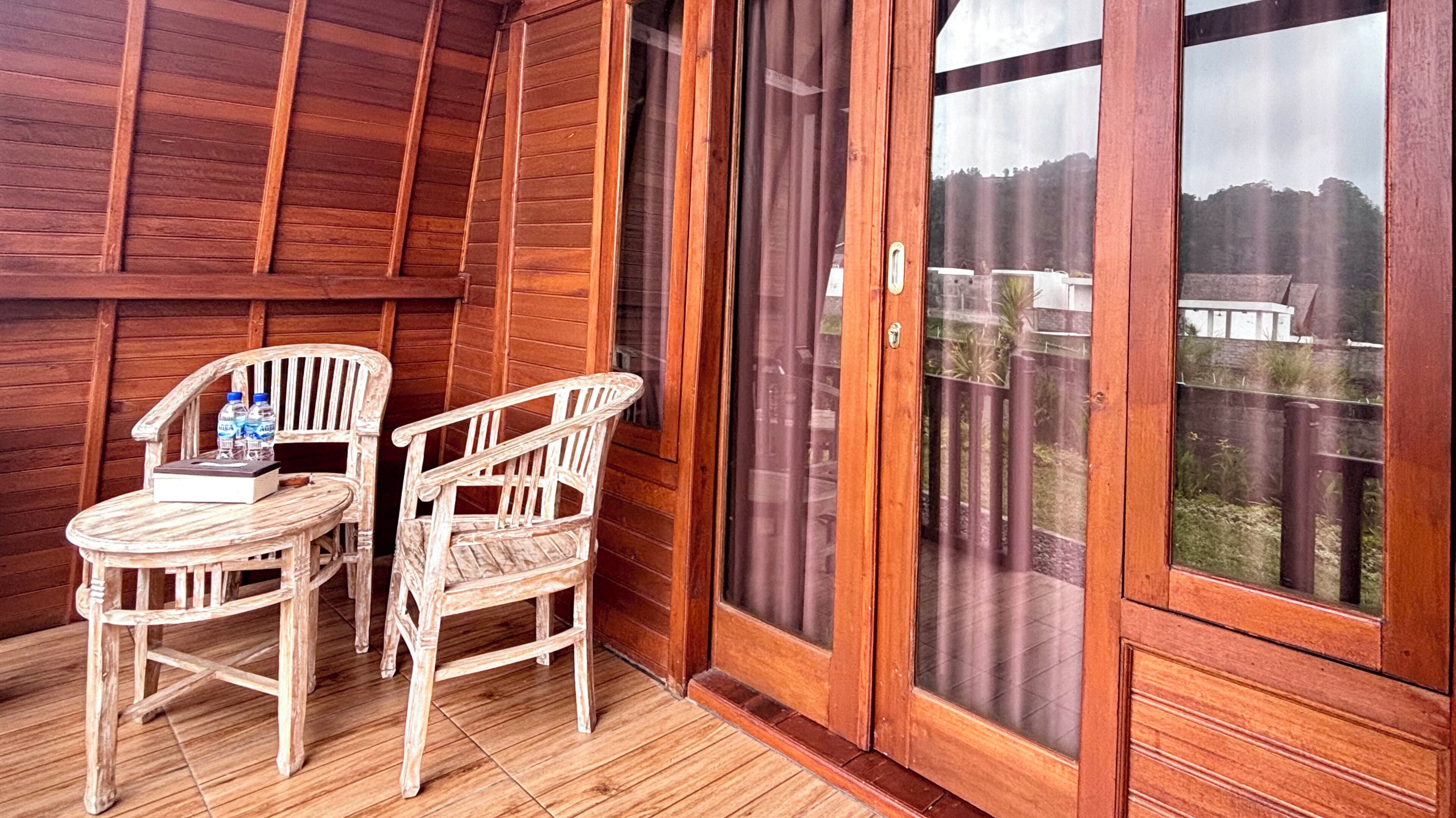 Een houten balkon met twee stoelen en een tafel, een schuifdeur van glas, en uitzicht op een hek en bomen.