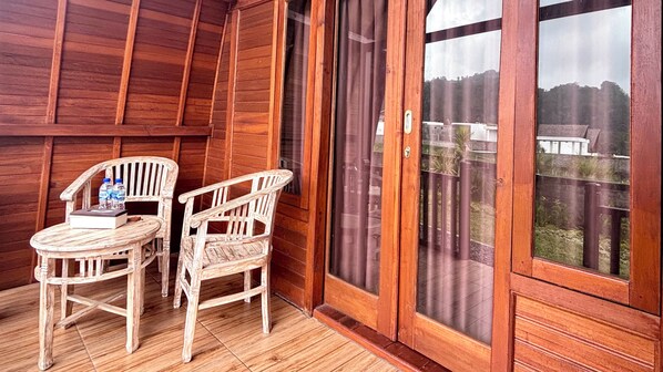 Un balcón de madera con dos sillas, una mesa, un ventanal corredizo y vistas a una valla y árboles.