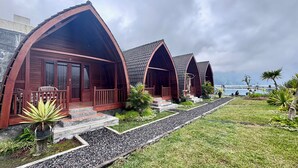 A row of wooden cabins with arched roofs, a gravel pathway, and a view of the sea. - Arkatama Lumbung Bali (Kintamani)