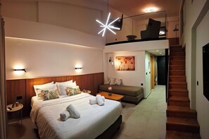 A modern hotel room with a large bed, a sofa, a small table, and a staircase leading to an upper level. - Aelia Luxury Apartments & Pool (Skafidia)