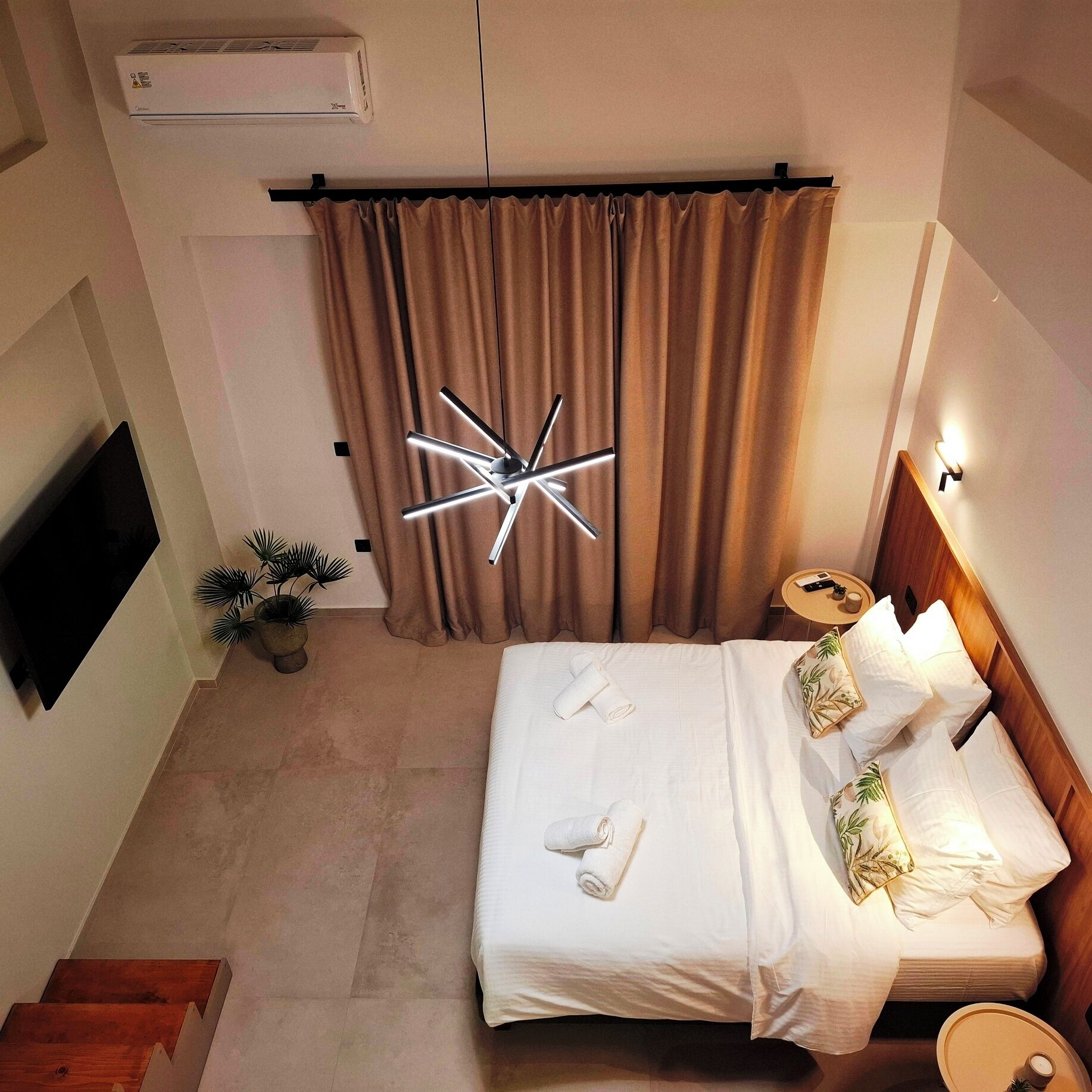 A hotel room with a bed, a TV, a wall-mounted air conditioner, a ceiling light fixture, and a small table with a remote control.