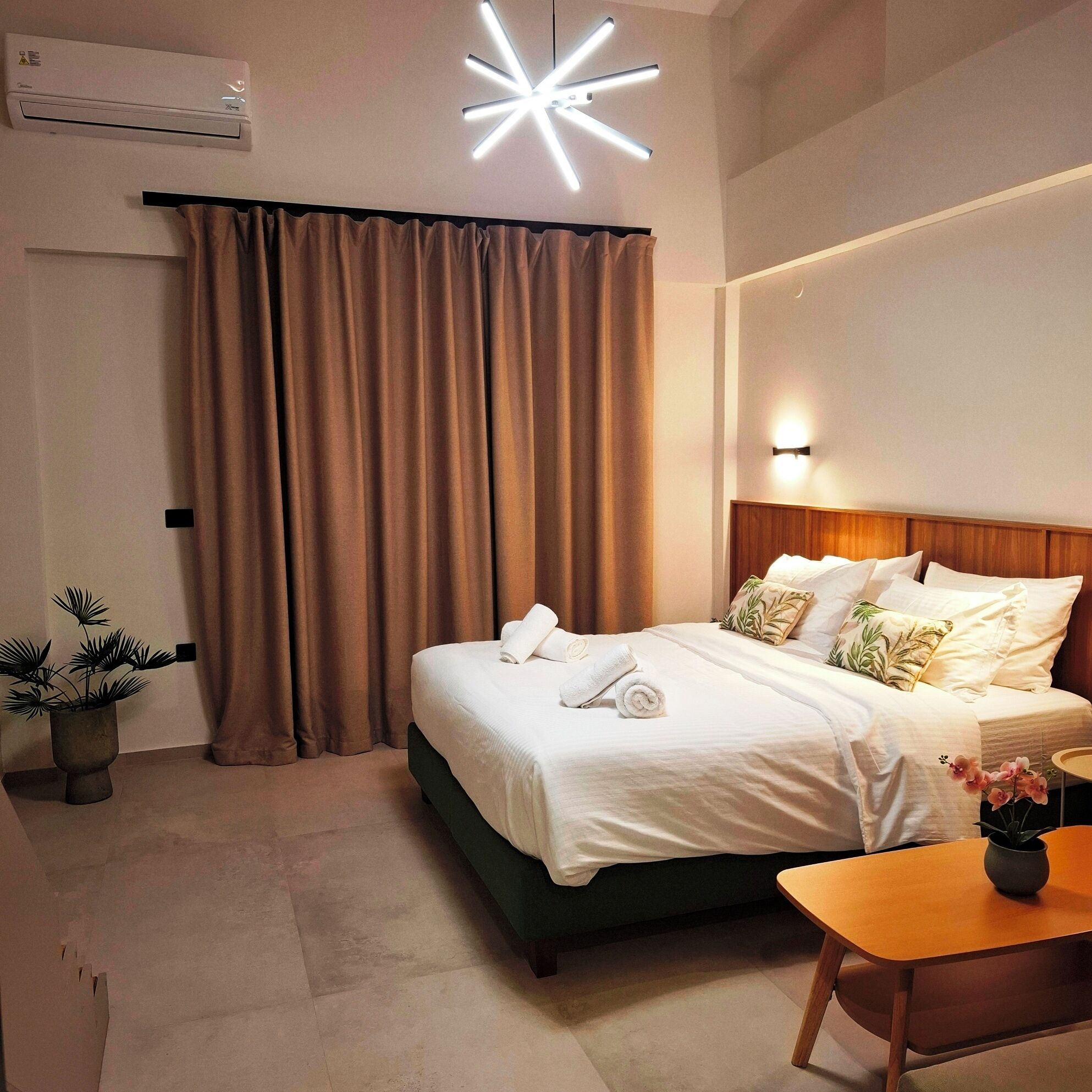A hotel room with a bed, a bedside table, a potted plant, a wall-mounted air conditioner, and a modern ceiling light fixture.