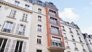 Un immeuble à plusieurs étages aux façades blanches et rouges, doté de nombreux balcons ornés de garde-corps en fer forgé, et surplombant un ciel dégagé.