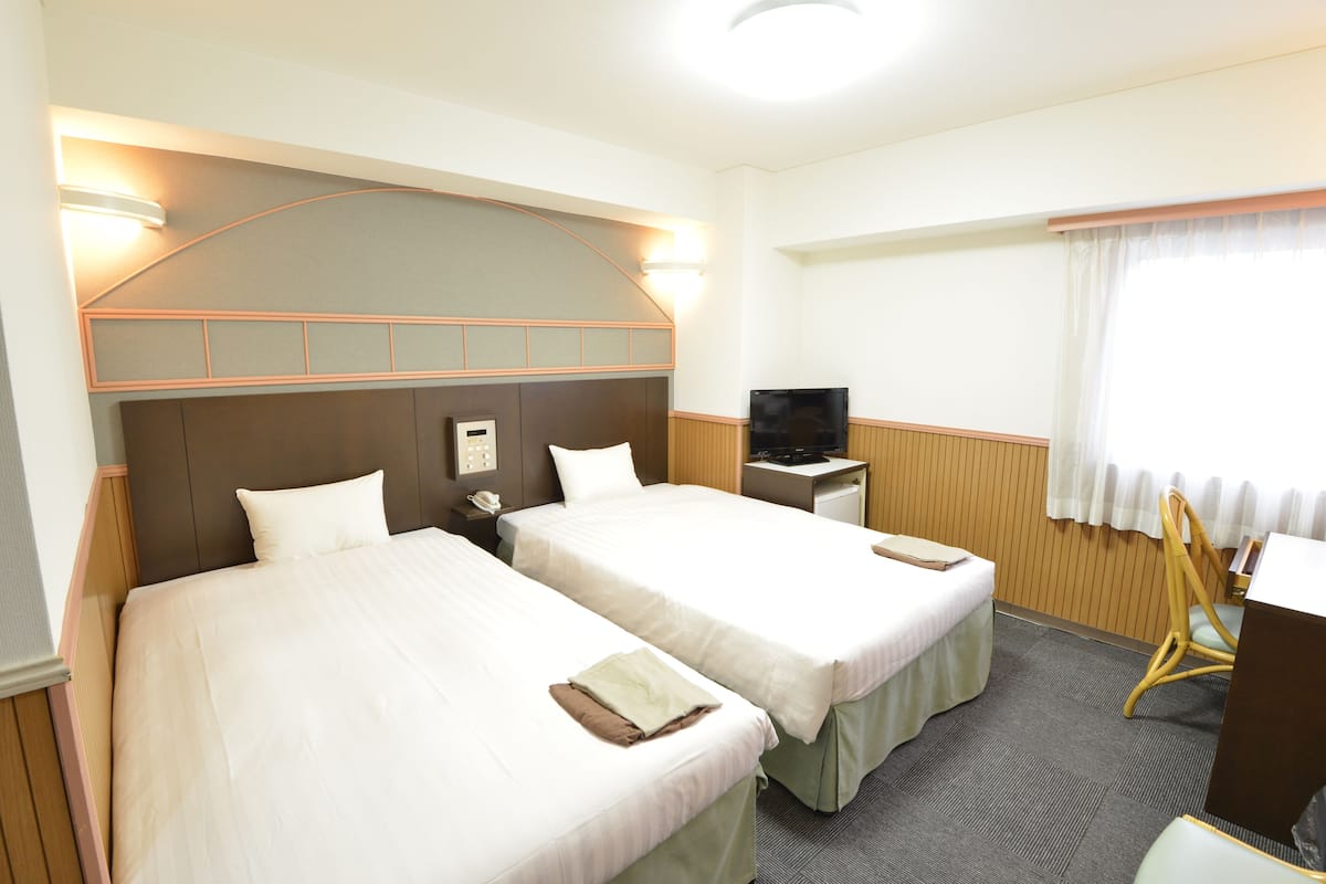 A hotel room with two beds, a desk, a television, and a window with curtains.