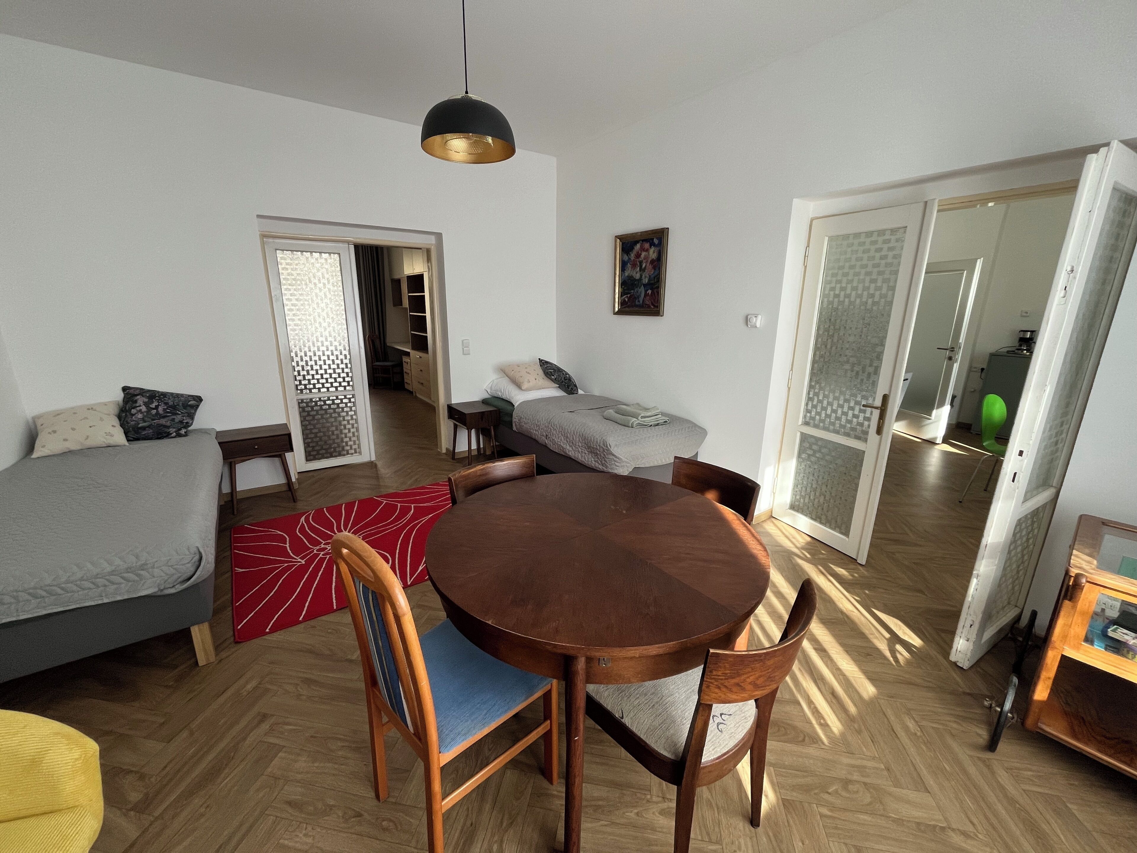 A modern living room with a round wooden table, two sofas, a dining table, and a large window.