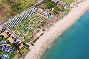 Ein Luftbild eines Küstenhotels mit einer langen Reihung von Zimmern, einem Strand und einem Poolbereich.