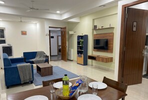 A living room with a sofa, coffee table, and a TV. There is a dining table with plates and glasses.