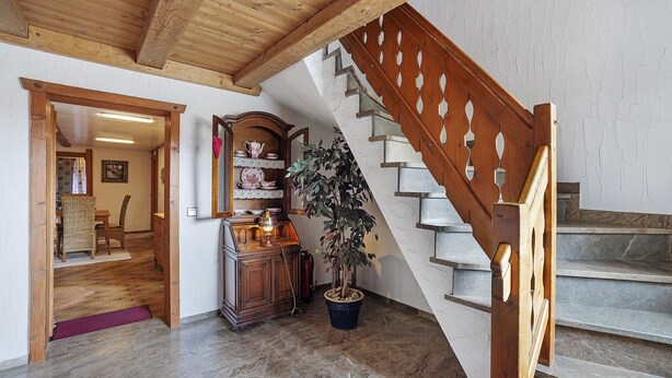 Un escalier en bois avec une rampe, une plante en pot, un meuble en bois orné d’objets décoratifs et un coin repas avec une table et des chaises.