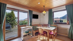 Une pièce chaleureuse avec un plafond en bois, une table à manger avec des chaises, un banc, un téléviseur fixé au mur et de grandes fenêtres avec des rideaux.