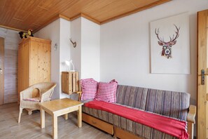 Ein gemütliches Zimmer mit Holzbalkendecke, einem Korbstuhl, einer Couch mit roten und weißen Karokissen und einem an der Wand befestigten Hirsch-Dekoration.