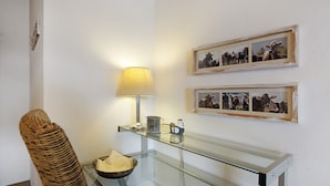 Une table avec un plateau en verre, une lampe, une chaise en osier et des tableaux encadrés accrochés au mur.