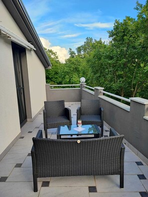 Ein Balkon mit Korbmöbeln und einem Glastisch, umgeben von einer Brüstung und Bäumen.