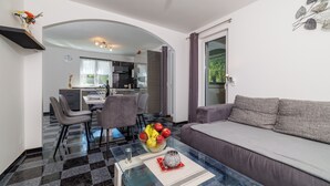 Ein modernes Wohnzimmer mit einem Glastisch, einem grauen Sofa und einem Essbereich mit Stühlen. Im Hintergrund ist eine Küche zu sehen.