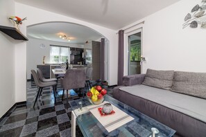 A modern living room with a glass coffee table, grey sofa, and a dining area with chairs and a kitchen in the background.