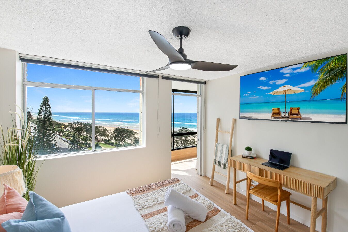 Ein Schlafzimmer mit großem Fenster, Deckenventilator, Schreibtisch und einem Gemälde mit Strandblick.