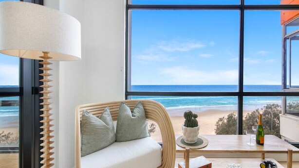 A room with a large window overlooking a beach, a wicker sofa with cushions, a small table with a bottle, glasses, and a book. - Pure Sunrise Oceanview 2BR Sub-Penthouse +Parking (Surfers Paradise)