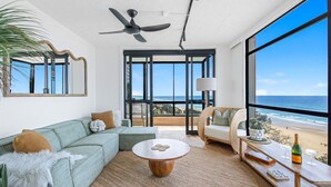 A spacious living room with a large window overlooking the beach, a ceiling fan, and a cozy seating area. - Pure Sunrise Oceanview 2BR Sub-Penthouse +Parking (Surfers Paradise)