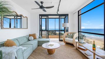 A spacious living room with a large window overlooking the beach, a ceiling fan, and a cozy seating area.