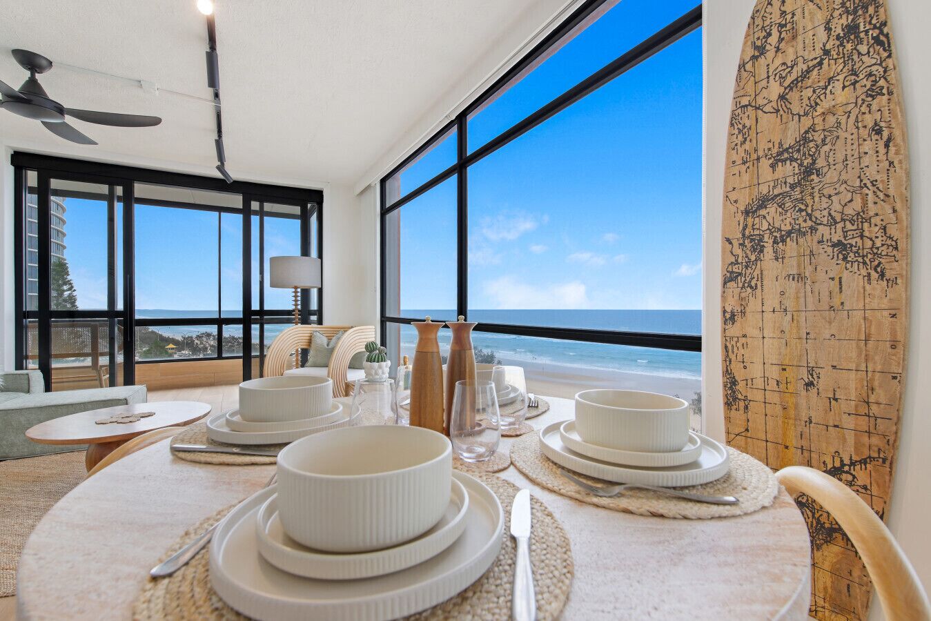 A dining area with a view of the beach, large windows, and a surfboard on the wall.