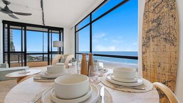 A dining area with a view of the beach, large windows, and a surfboard on the wall.