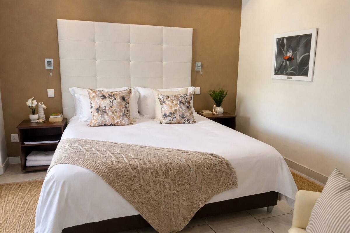 A neatly made bed with a white duvet and two decorative pillows, a wooden nightstand with a plant, and a framed picture on the wall.
