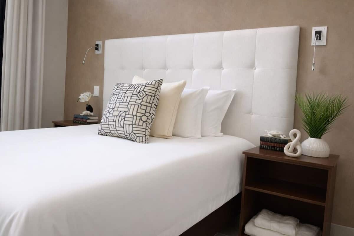 A neatly made bed with a tufted headboard, a patterned pillow, and a bedside table with a plant and books.