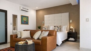 A modern hotel room with a brown leather sofa, a round black ottoman, a large bed with a tufted headboard, and a nightstand with a vase of flowers. - Monte Bello Guesthouse (Windhoek)
