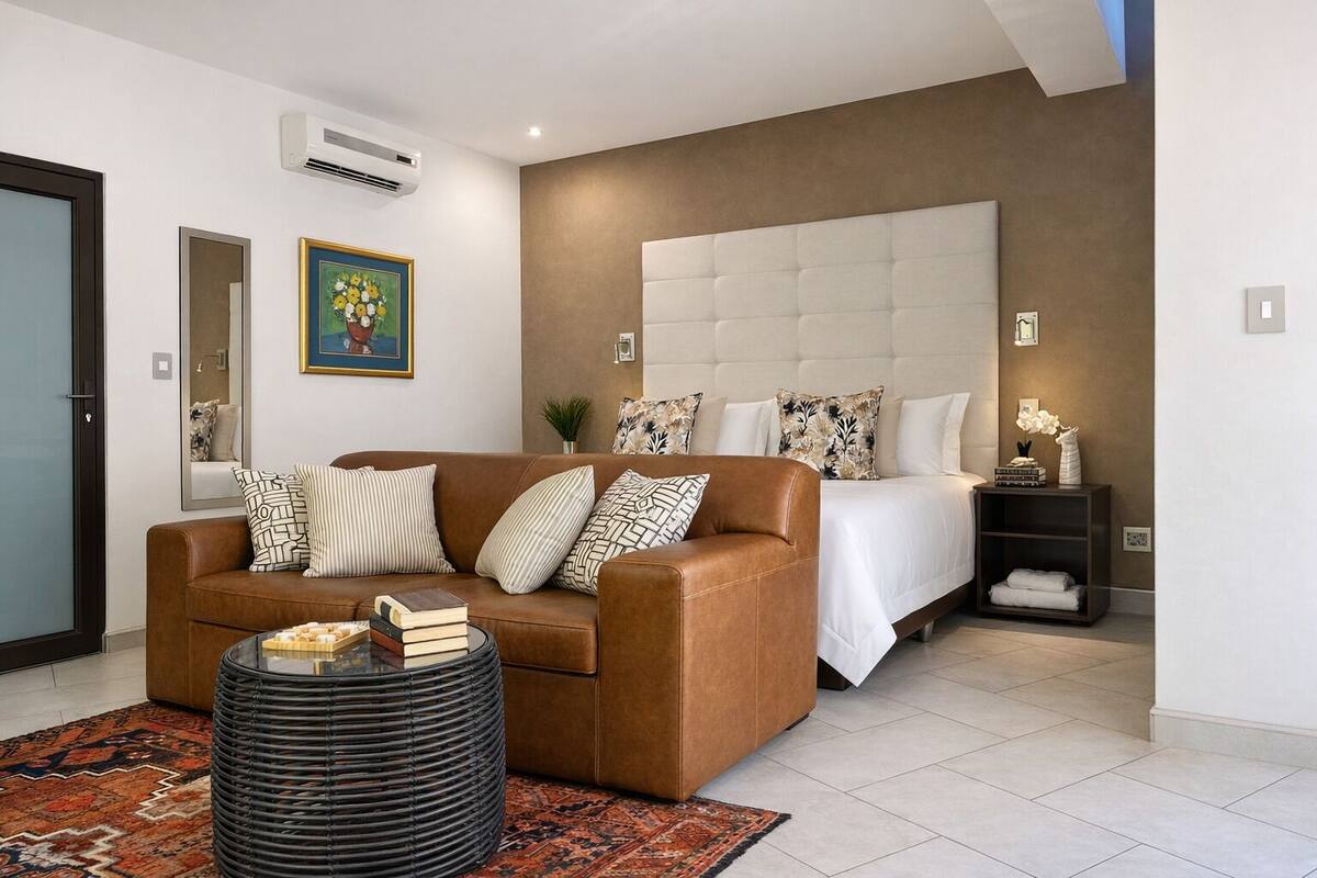 A modern hotel room with a brown leather sofa, a round black ottoman, a large bed with a tufted headboard, and a nightstand with a vase of flowers.