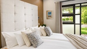 A hotel room with a large bed, multiple pillows, a headboard, a window with curtains, and a framed picture on the wall. - Monte Bello Guesthouse (Windhoek)