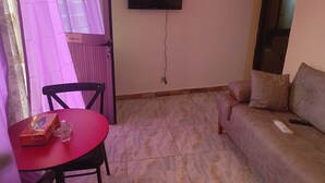 Ein Wohnzimmer mit einem roten Tisch, einem schwarzen Sessel, einem beigen Sofa und einem Wand-Fernseher.