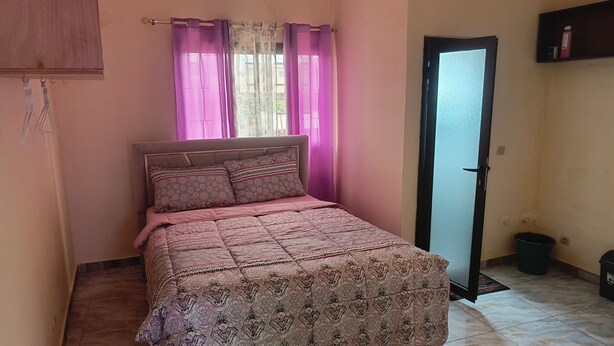 A bedroom with a bed, curtains, an air conditioning unit, a door, and a shelf. - BEL APPARTEMENT 1-Likouala! A stone's throw from downtown (LIBREVILLE)