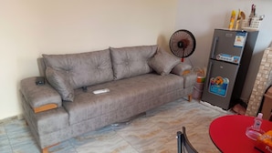 A living room with a sofa, a fan, a refrigerator, and a dining table. - BEL APPARTEMENT 1-Likouala! A stone's throw from downtown (LIBREVILLE)