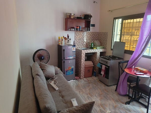 A compact living space with a sofa, kitchenette, fan, and a window with curtains.